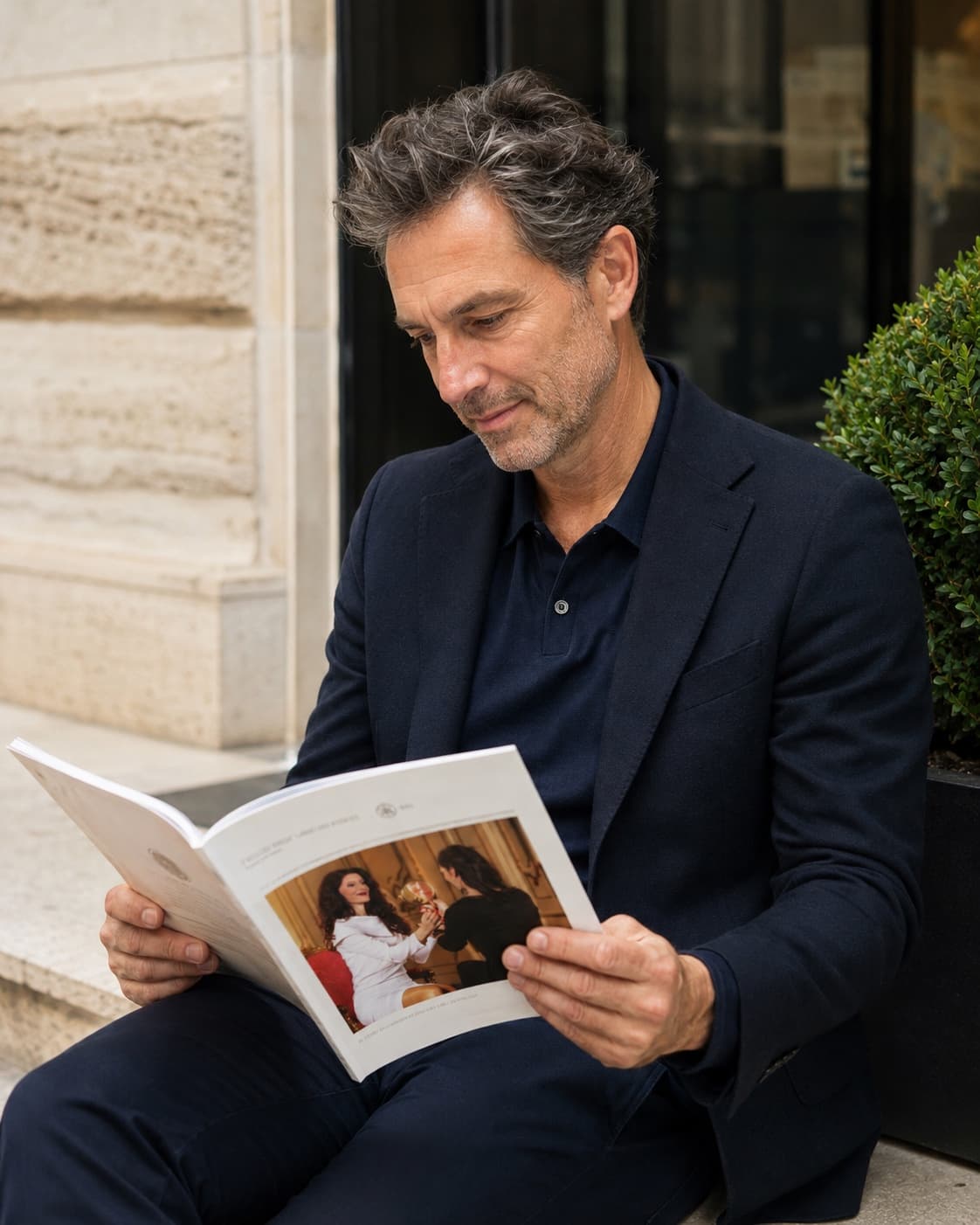 Gepflegter Mann um die 50 im dunkelblauen Sakko liest entspannt ein Magazin im hellen Tageslicht vor einer klassischen Fassade – Sinnbild für datenbasierte Longevity-Medizin für Männer. Model, kein Patient.